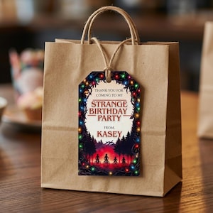May include: A brown paper gift bag with a tag that reads "Thank you for coming to my Strange Birthday Party from Kasey." The tag features a colourful illustration of a forest scene with silhouettes of people on bikes.