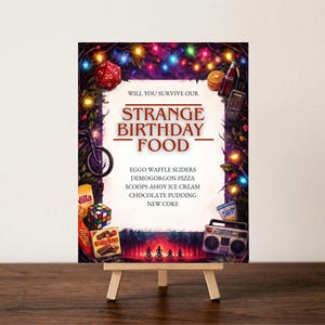 May include: A sign on a small easel with the words "STRANGE BIRTHDAY FOOD". The sign features a red D20 die, a Rubik's Cube, and a retro boombox. The food list includes Eggo Waffle Sliders and Demogorgon Pizza.