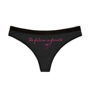 May include: Black underwear with a black waistband. The phrase "the future is female" is written in pink script across the front, with a pink bow graphic underneath. The underwear is a thong style.