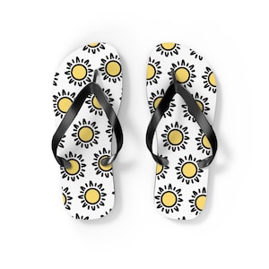 Op de afbeelding: Een paar witte teenslippers met een herhaald patroon van gele zonnen met zwarte contouren. De teenslippers hebben zwarte bandjes en zijn ontworpen voor casual gebruik. Het ontwerp is vrolijk en geschikt voor de zomer.