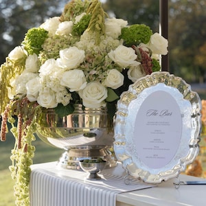 May include: A bar display featuring a large silver bowl filled with white roses and green hydrangeas. A silver tray with a menu reading "The Bar" is on a white table with a striped runner.
