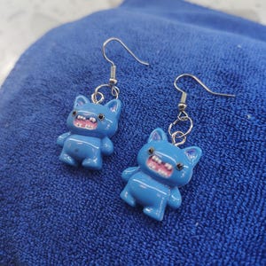 May include: A pair of blue cat-shaped earrings with silver-colored ear wires. Each earring features a cartoon cat with a wide, toothy grin and small arms and legs. The earrings are set against a blue background.