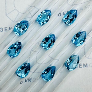 Op de afbeelding: Negen peervormige, gefacetteerde, lichtblauwe edelstenen op een witte ondergrond. De edelstenen zijn geslepen om licht te reflecteren, waardoor hun interne structuur zichtbaar wordt. Waarschijnlijk aquamarijn of topaas.