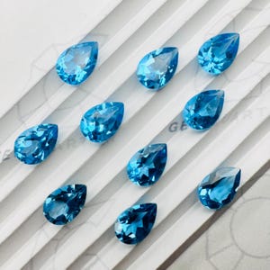 以下が含まれることがあります： 鮮やかなクリアブルーの色合いの、12個のペアシェイプのファセット宝石のコレクション。 宝石は、視覚的に魅力的なディスプレイを作成する、微妙な線状パターンを持つ白い表面に配置されています。 宝石はジュエリー作りのためです。
