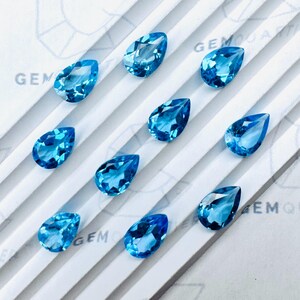 以下が含まれることがあります： さまざまな青色の、12個のペアシェイプのファセット宝石のコレクション。宝石は、斜めの線が入った白い表面に配置されています。「GEM」という単語が背景に部分的に表示されています。各宝石は約1cmです。