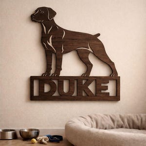 May include: A wooden wall art piece featuring a dog silhouette with the name "DUKE" cut out below. The dog is a dark brown color, and the name is in a block font. The art is mounted on a light-colored wall.