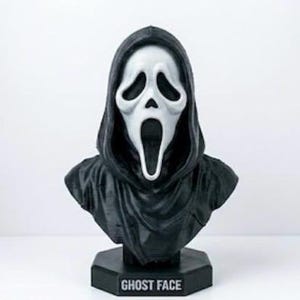 Könnte beinhalten: Eine Ghost Face Büste mit einer weißen Maske, länglichen schwarzen Augenhöhlen und einem weit geöffneten Mund, gehüllt in eine schwarze Kapuzenrobe. Die Figur steht auf einem schwarzen sechseckigen Sockel mit dem Schriftzug GHOST FACE in Weiß.