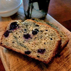 Puede incluir: Dos rebanadas de pan de arándanos en una tabla de madera. El pan tiene una corteza dorada y está salpicado de arándanos azul oscuro. Un cuenco blanco y una taza decorativa están en el fondo.