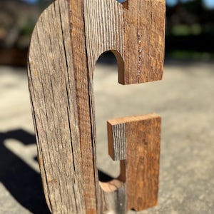 May include: A large wooden letter "G" stands upright, crafted from reclaimed wood. The letter displays a mix of light and dark brown tones, with visible wood grain and texture. The rustic design is suitable for home decor.