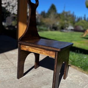 May include: A handcrafted wooden chair with a dark stain finish. The chair features a high back with a handle, a rectangular seat, and arched legs. The chair's dimensions are approximately 15 inches wide, 12 inches deep, and 30 inches tall.
