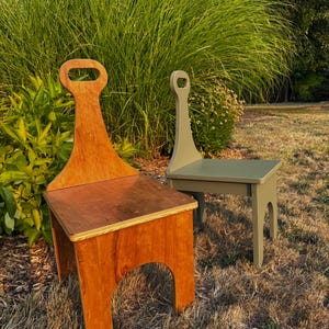 May include: Two wooden chairs with high backrests and integrated handle cutouts. One chair features a natural wood grain finish with visible plywood edges, while the second chair is painted in a solid muted olive green color.