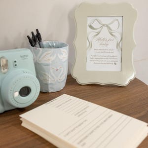 May include: A light blue Instax Mini camera is next to a swan-patterned pen holder and a framed card reading "Notes for baby." A stack of lined cards is in the foreground. The items are on a wooden surface.