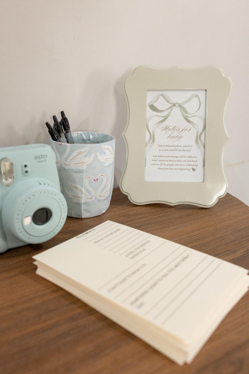 May include: A light blue Instax Mini camera is next to a swan-patterned pen holder and a framed card reading "Notes for baby." A stack of lined cards is in the foreground. The items are on a wooden surface.