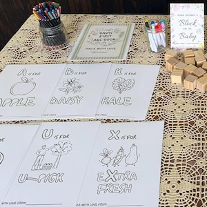 May include: A baby shower activity set featuring printed coloring sheets with letters and illustrations, a jar of colored pencils, a jar of markers, a sign reading "Please decorate a block for the baby," and a pile of small, unfinished wooden blocks.