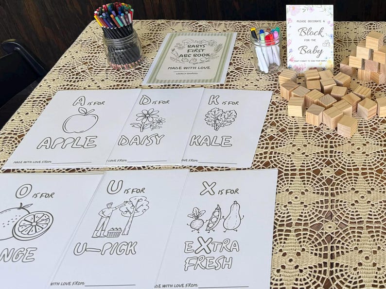 May include: A baby shower activity set featuring printed coloring sheets with letters and illustrations, a jar of colored pencils, a jar of markers, a sign reading "Please decorate a block for the baby," and a pile of small, unfinished wooden blocks.