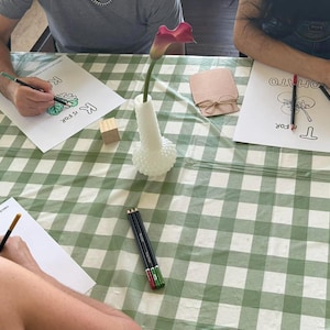 May include: Three people coloring alphabet activity sheets with colored pencils on a green and white checkered tablecloth. A white hobnail vase holds a single purple calla lily, and a small wooden block sits near a brown paper cutout jar.