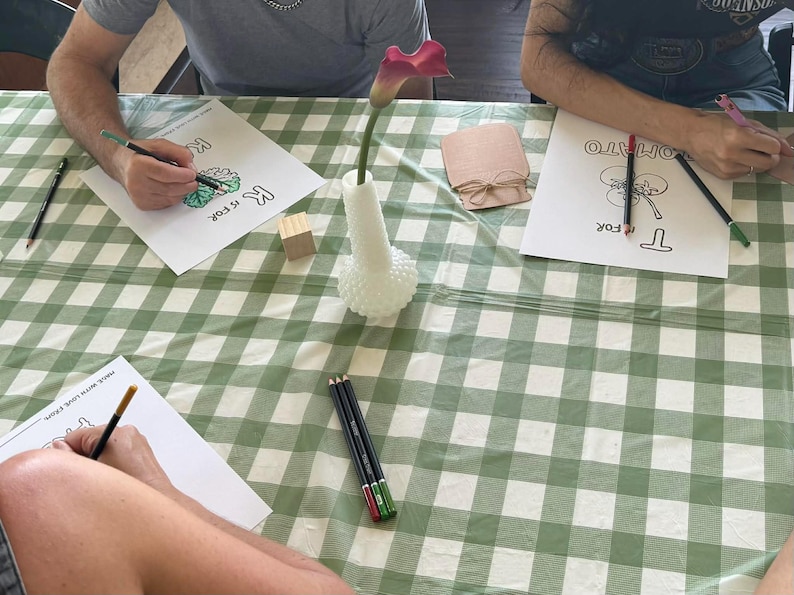 May include: Three people coloring alphabet activity sheets with colored pencils on a green and white checkered tablecloth. A white hobnail vase holds a single purple calla lily, and a small wooden block sits near a brown paper cutout jar.