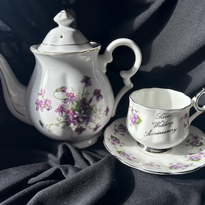 May include: A white porcelain tea set featuring a teapot, teacup, and saucer. The set is decorated with purple floral patterns and gold trim. The teacup and saucer have the text "Silver Wedding Anniversary".