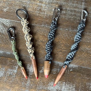 May include: Four bullet-shaped keychains with braided cord and metal clasps. The bullet casings are copper-coloured with red tips. The cords are in shades of green, tan, and grey. One keychain has a keyring.
