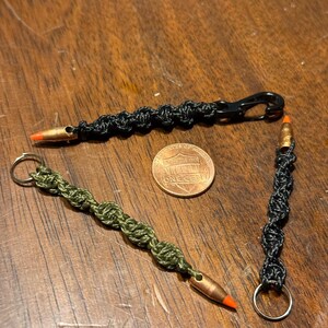 May include: Three handmade keychains with bullet casings and knotted cord. One keychain is black with a black clasp, another is olive green with a silver key ring, and the third is black with a silver key ring. A penny is also in the image.