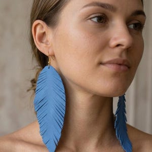 Peut inclure: Une paire de grandes boucles d'oreilles bleues en forme de plume avec un bord frangé. Chaque boucle présente une surface texturée et est suspendue à un crochet en métal doré, créant une silhouette longue et pendante qui atteint le cou.