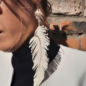 May include: A close-up of a white feather earring. The earring is long and detailed, with individual feather segments. The person is wearing a black turtleneck and a white blazer. The background is a brick wall.