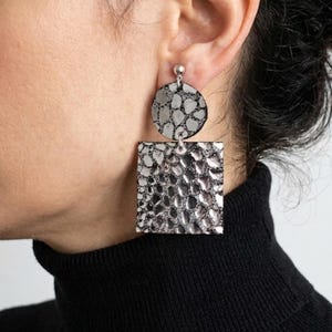 May include: Silver earrings with a textured, reptile-like pattern. Each earring has a circular top and a square bottom, connected by a small silver loop. The earrings are shown against a black turtleneck.