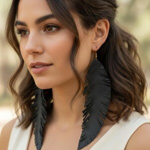 May include: Black feather earrings with gold-colored hooks. The earrings are long and dangle, with a detailed feather design. The earrings are worn by a person with brown hair and a white top.