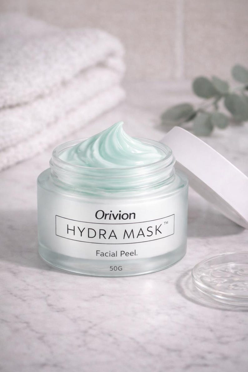 May include: A jar of Orivion Hydra Mask facial peel with a light blue, creamy product. The jar is open, with the lid resting beside it. The label reads "Hydra Mask Facial Peel" and "50G". A white towel is in the background.