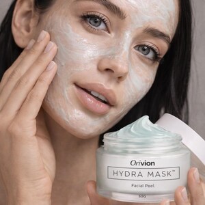 May include: A person with a facial mask applied, holding a jar of Orivion Hydra Mask. The jar is clear with a white label and a light blue cream. The text "Hydra Mask" and "Facial Peel" are visible on the label. The jar contains 50g of product.