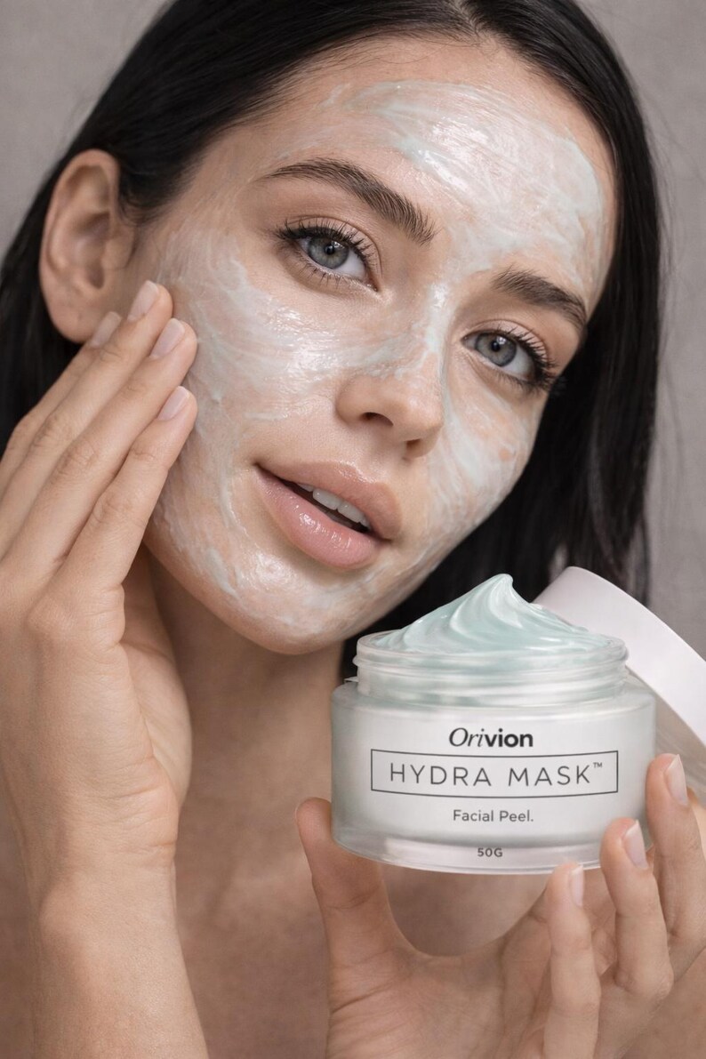 May include: A person with a facial mask applied, holding a jar of Orivion Hydra Mask. The jar is clear with a white label and a light blue cream. The text "Hydra Mask" and "Facial Peel" are visible on the label. The jar contains 50g of product.