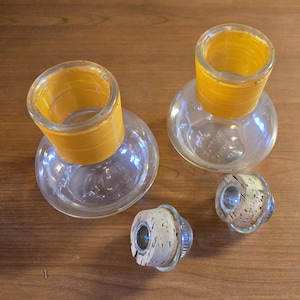 May include: Clear glass bottles with yellow rims and cork stoppers. The bottles have a rounded base and a cylindrical neck. The cork stoppers are made of cork and have a metal ring around them. The bottles and stoppers are on a wooden surface.