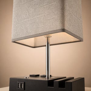 May include: A modern table lamp with a rectangular gray fabric shade and a chrome-colored metal stem. The black base features two USB ports and slots for devices. The lamp is designed for bedside or desk use.