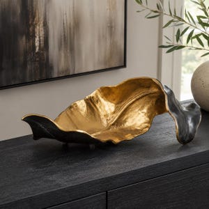 May include: A decorative bowl shaped like a large, elongated leaf. The interior surface features a bright, metallic gold finish with a textured, organic appearance, while the exterior is finished in a contrasting matte black color.