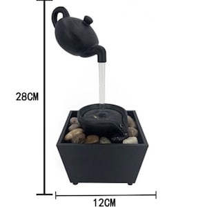 May include: A black tabletop water fountain with a teapot design. The fountain features a teapot pouring water into a small basin, which then flows into a square base filled with stones. The fountain is 11 inches tall and 4.7 inches wide.