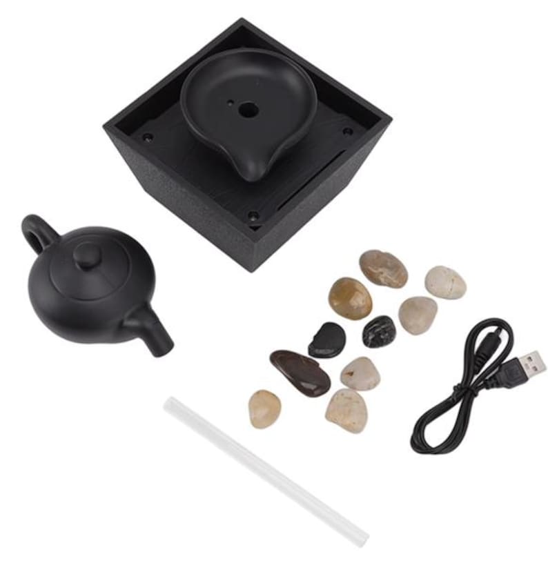 May include: A black tabletop fountain kit with a small teapot, a square base with a water basin, smooth stones, a clear straw, and a USB cable. The fountain base has a unique, modern design.