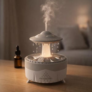 May include: A white and gray essential oil diffuser with a unique UFO-like design. The diffuser emits a stream of vapor and has a glowing light. A small brown bottle with a dropper is visible in the background.
