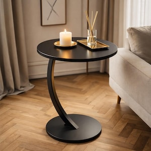 May include: A black side table with a round top and a curved leg. A lit candle sits on a gold-colored plate, and a tray with reeds and a glass of water are on the table. The table is on a wooden floor.