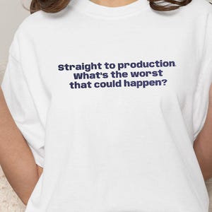 May include: White t-shirt with the text "Straight to production. What's the worst that could happen?" in navy blue. The person is wearing denim shorts and white socks with black stripes.