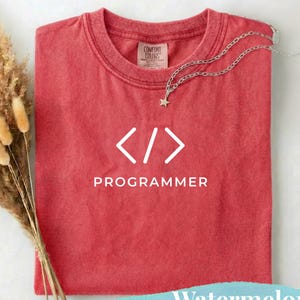 Könnte beinhalten: Ein rotes T-Shirt mit dem Wort "PROGRAMMER" und Codesymbolen in Weiß gedruckt. Eine silberne Halskette mit einem Sternanhänger liegt auf dem Shirt. Das Shirt ist gefaltet und hat ein "Comfort Colors"-Etikett.