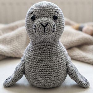 Puede incluir: Un peluche de foca gris hecho a mano con ganchillo, con ojos de botón negros y una nariz y bigotes bordados en negro. La foca tiene pequeñas aletas y está sentada erguida. El juguete está hecho de hilo suave.
