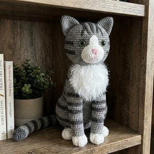Könnte beinhalten: Eine handgefertigte Häkelkatzenpuppe in Grautönen mit schwarzen Streifen, weißen Akzenten und grünen Augen. Die Katze hat einen flauschigen weißen Kragen und sitzt auf einem Holzregal. Bücher und eine kleine Topfpflanze befinden sich im Hintergrund.
