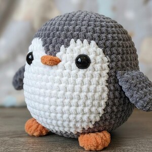 Può includere: Un peluche a forma di pinguino fatto a mano all'uncinetto. Il pinguino ha la testa e le ali grigie, la pancia bianca e le zampe e il becco arancioni. Ha grandi occhi neri. Il peluche è rotondo e morbido, perfetto come giocattolo per bambini o per l'arredamento della cameretta.