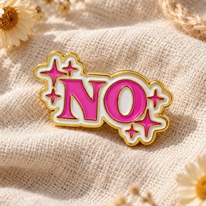 Pode incluir: Um broche de esmalte com borda dourada apresentando a palavra NO em letras rosa brilhante sobre um fundo branco. O design é acentuado com várias pequenas estrelas rosa de quatro pontas que cercam o texto, criando um visual marcante.