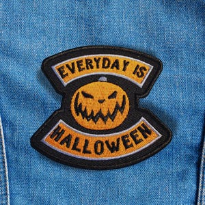 Może przedstawiać: Haftowana naszywka z napisem "EVERYDAY IS HALLOWEEN" w żółtych literach na czarnym tle. Naszywka przedstawia wzór dyni na Halloween w kolorze pomarańczowym i czarnym, na niebieskim tle z dżinsu.
