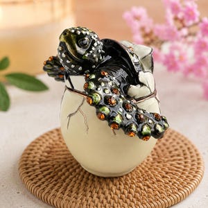 Può includere: Un portagioie decorativo a forma di tartaruga marina nera che esce da un uovo color crema. La tartaruga presenta smalto scuro con accenti verdi e piccoli cristalli arancioni incastonati lungo il guscio e le pinne. L'uovo ha un design crepato.