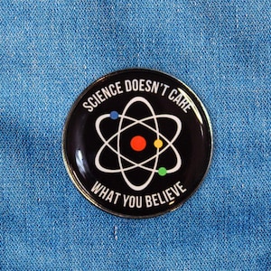 Peut inclure: Un badge rond noir avec le texte "SCIENCE DOESN'T CARE WHAT YOU BELIEVE" en blanc. Il présente un symbole d'atome blanc avec des accents rouges, jaunes et bleus. Le badge est épinglé sur une veste en jean bleue.