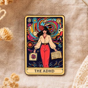 Könnte beinhalten: Eine rechteckige Emaille-Anstecknadel mit einem Tarotkarten-Design, das eine zentrale Figur in einem cremefarbenen Hemd und roter Hose zeigt, umgeben von bunten Wirbelmustern, verschiedenen Symbolen und dem Text THE ADHD auf einem Banner unten.