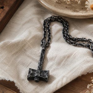 Könnte beinhalten: Eine silberfarbene Anhängerkette mit einem hammerförmigen Anhänger. Der Anhänger hat ein strukturiertes, gehämmertes Aussehen und hängt an einer passenden Kette. Die Halskette liegt auf einem cremefarbenen Tuch, mit einem Räucherstäbchenhalter aus Holz und Trockenblumen im Hintergrund.