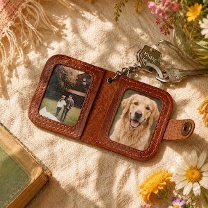 Op de afbeelding: Een bruine leren sleutelhangerportemonnee met twee fotovensters, één met een stel en de andere met een golden retriever. De portemonnee is open en toont de foto's, bevestigd aan sleutels. De achtergrond omvat bloemen en een vintage boek.